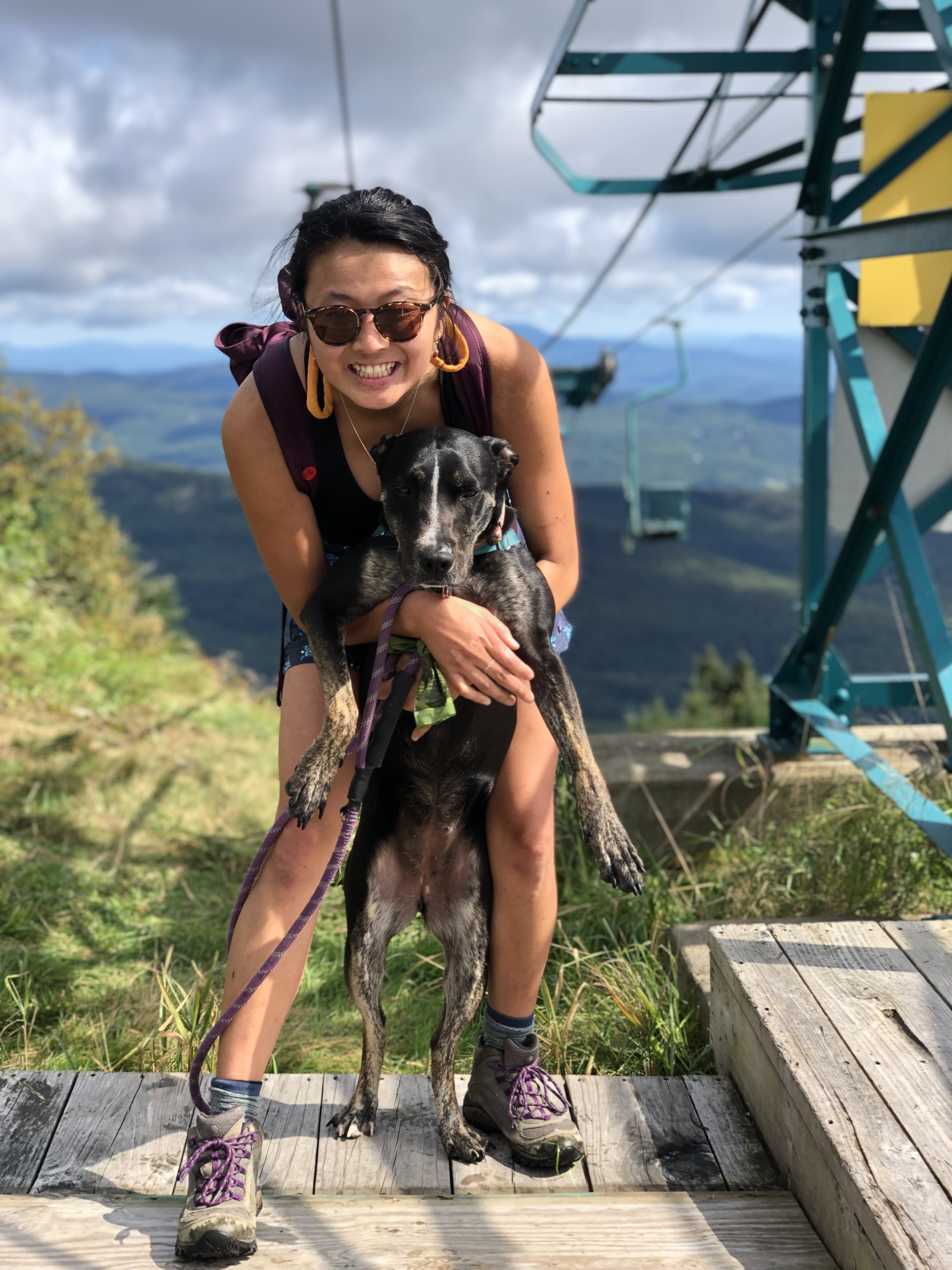 A women with dark hair wearing sunglasses is holding a black dog with a view of green trees and a ski lift in the background