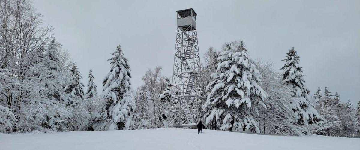 A winter hiker snowshoes up to a very snowy firetower at Allis State Park.